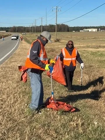 Two volunteers in safety vests collecting litter during a roadside cleanup