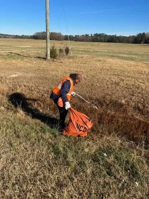Volunteer in safety gear collecting litter beside a rural roadside