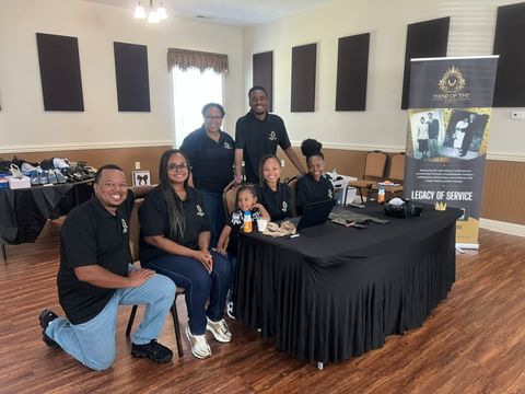 Hand of the King team gathered at a table beside the Legacy of Service banner at the Clothing Giveaway
